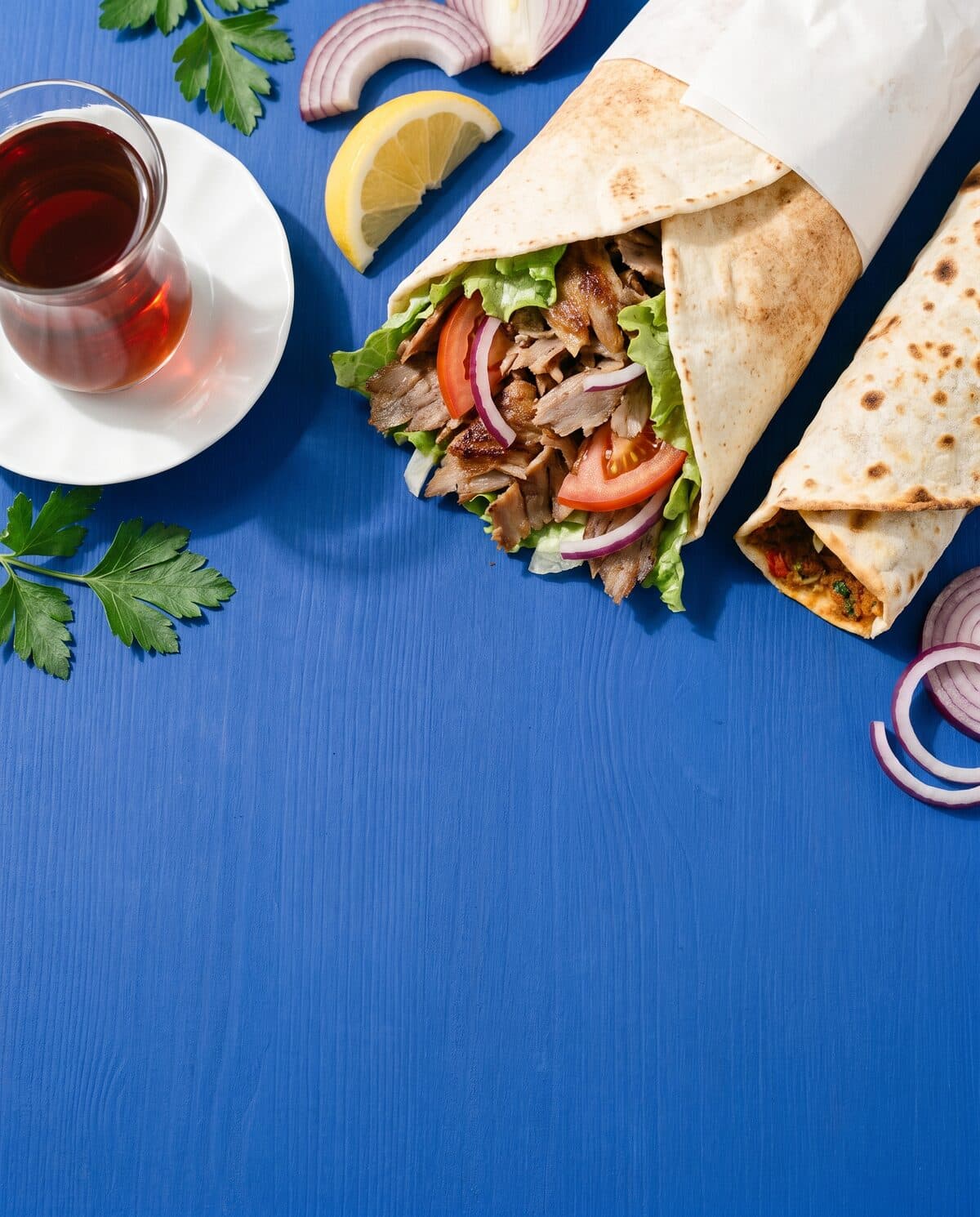 Beispielbild: Döner im Papier, gerollter Lahmacun, Zitrone und Tee im Glas von oben auf kobaltblauer Fläche