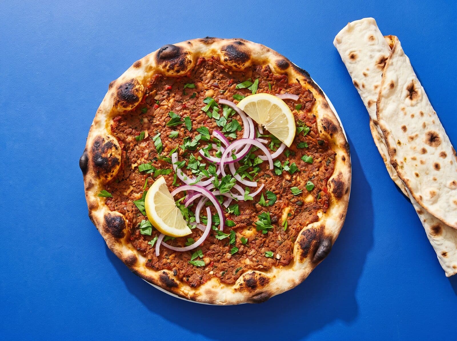 Beispielbild: Lahmacun mit Petersilie, roten Zwiebelringen und Zitronenstücken von oben auf blauer Fläche, daneben gerolltes Fladenbrot