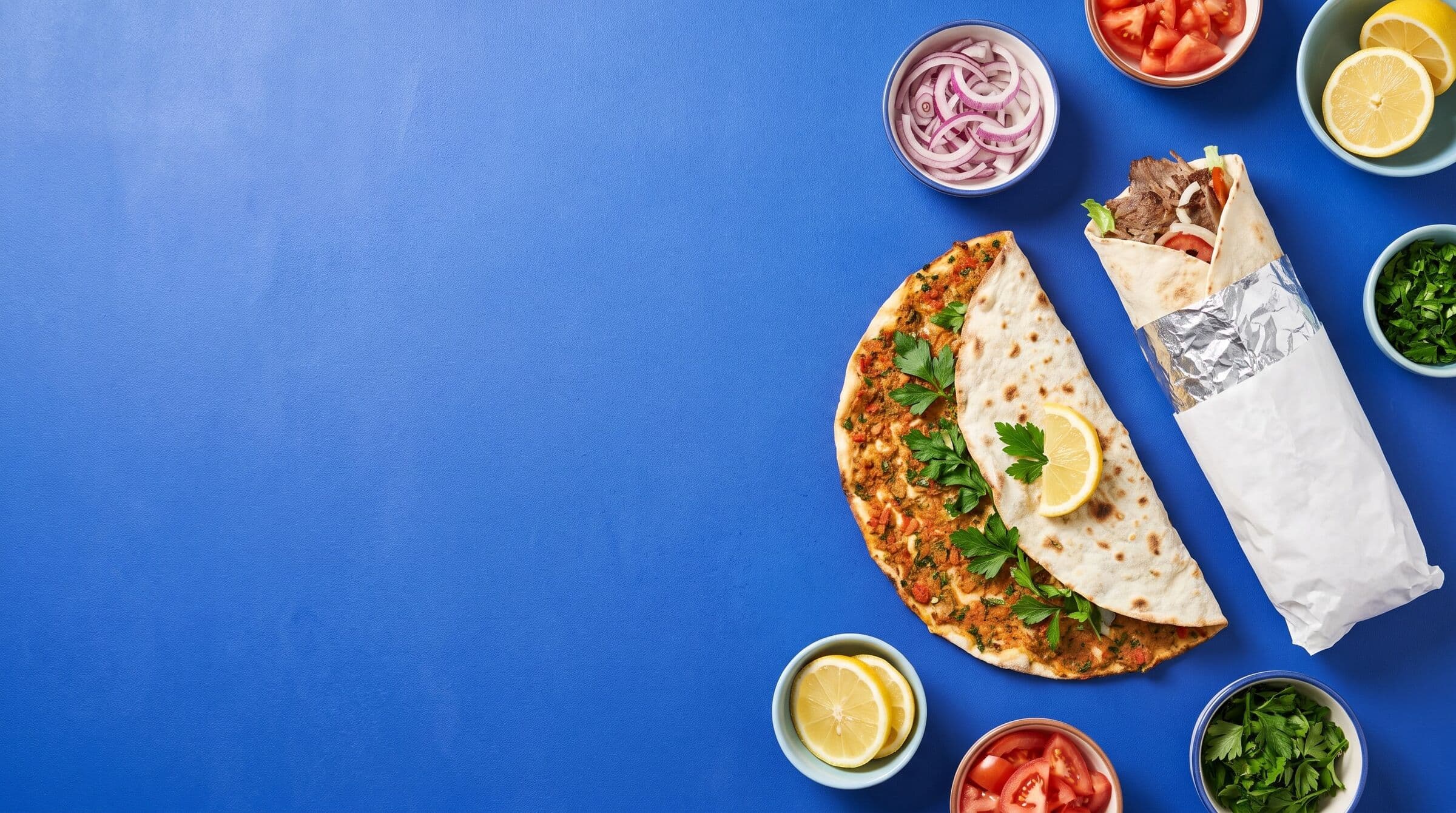 Beispielbild: Halb zugeklappter Lahmacun, Döner in Folie und Papier und Schälchen mit Zwiebeln, Tomaten und Petersilie auf blauer Fläche